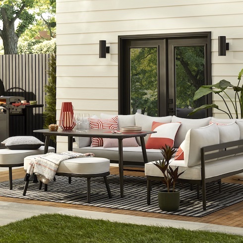 Beautiful backyard wood patio eating area featuring The Home Depot’s Hampton Bay Claxton Steel 5-Piece Patio Sectional Dining Set and Grey Stripe Indoor/Outdoor Area Rug, two potted plants, white and red throw pillows, a woven throw, a white boarded exterior wall with black double doors and two cylindrical exterior lights, a barbeque, and a patch of grass