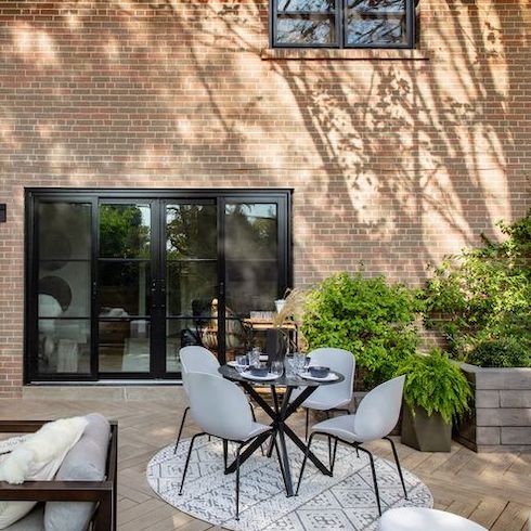 Outdoor area for al fresco dining with a round dining table, four grey plastic dining chairs, a large sliding black framed door and window set into a tall exterior brick wall with stamped pavers in a herringbone pattern on the ground, potted trees and ferns, and a raised brick garden bed with greenery