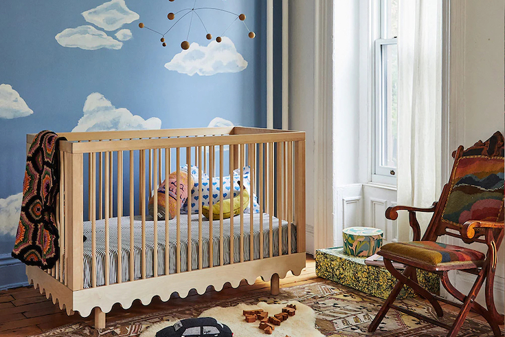 The Oeuf Moss Crib from Oeuf Canada, which features a bold-yet-simple wave motif and airy round spindles, sits in a charming modern nursery with a blue sky and white cloud wall mural, a modern metal and wood mobile overhead, a tall window with white gauzy drapes, two colourful boxes under the window, a brown and cream rug with a sheepskin rug on top, wooden floor, and an antique wood chair with colourful upholstery as part of HGTV.ca Nursery Trends for 2025