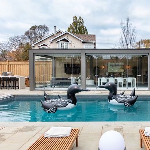A renovated backyard featured on HGTV Canada’s Backyard Builds featuring large sand patio stones, an inground pool with two blow up loon floaties, an eat at bar, and a covered outdoor kitchen with retractable glass walls by Lumon