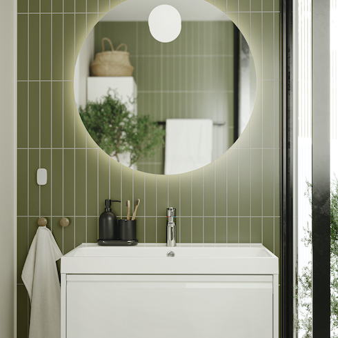 Sleek modern ÄNGSJÖN bathroom system from Ikea with a white vanity and sink, a large round backlit mirror, a black toothbrush set, two round towel hooks, and sage green subway tiles installed vertically shown in the HGTV Canada gallery Unusual Bathroom Renovation Ideas That Will Be Popular In 2025