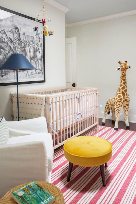 A chic circus themed nursery in Toronto designed by Kelly Lynn Armstrong featuring crisp white walls, black-and-white artwork, a tall super fun giraffe toy, a Restoration Hardware stool, a wooden crib, a navy blue floor lamp, a white nursing armchair, a small round wicker side table, and a red a white striped rug