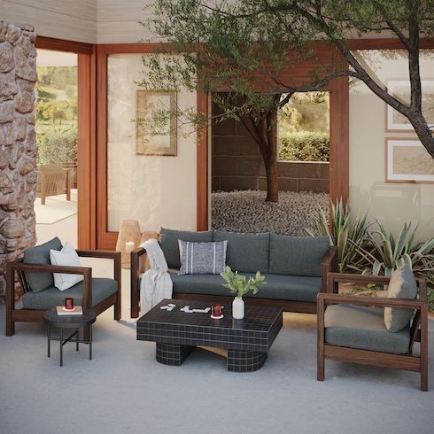 Walled outdoor courtyard with Palmera Lounge Chairs and Sofa, Kera black ceramic coffee table, and round side table all by Article, an olive tree, two potted aloes, and a doorway looking out over a rolling hill view