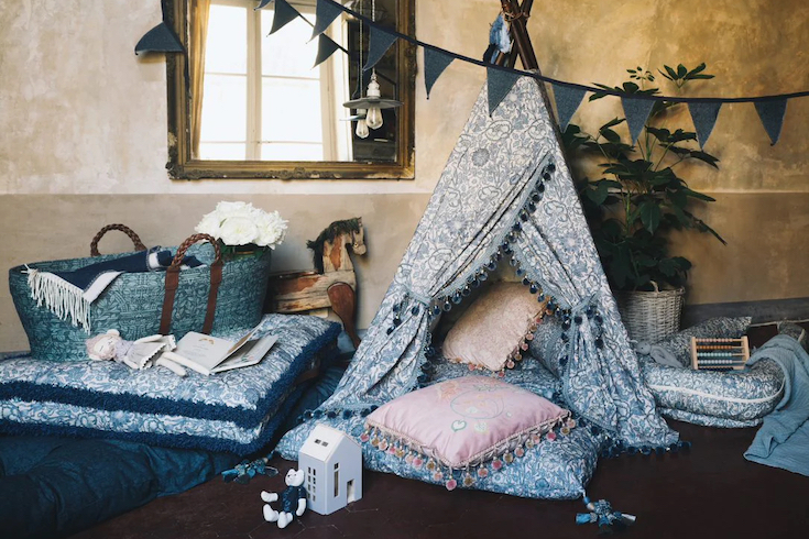 A romantic nursery shot by influencer and photographer Jamie Beck for DockATot x William Morris & Co. featuring floor cushions and a play tent in the Pink & Rose print, an antique rocking horse, blue bunting, blue market basket and toy house and bear on the floor
