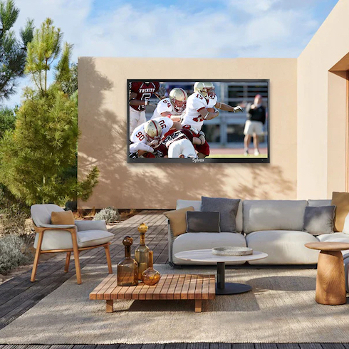 Sylvox’s waterproof, anti-glare outdoor TV with a football game playing hangs on the wall of an outdoor seating area of an adobe desert house with a sliding wall door, white sectional outdoor couch and armchair, and three outdoor side tables all sitting on a wooden patio next to a cactus and pine tree garden