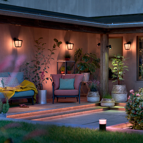 Wall sconces and path lights using Philips Hue Smart Lighting system are seen light up at dusk in a sunken outdoor courtyard, casting soft colourful light over an outdoor sofa and armchair under a covered porch, with multiple potted plants placed around and a grass lawn