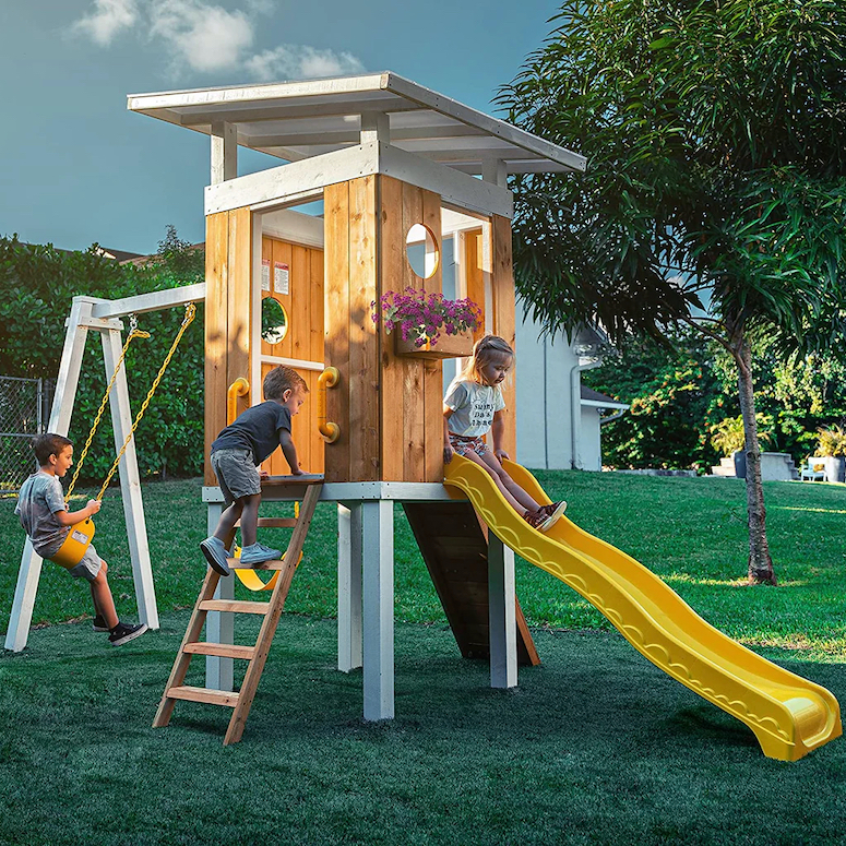 Avenlur Modern Backyard Outdoor Swing Set sold in Canada by WoodWood Toys sits in a green backyard at dust with three children playing on it as featured in Outdoor Home Playground Sets That Are Worth the Money for HGTV.ca