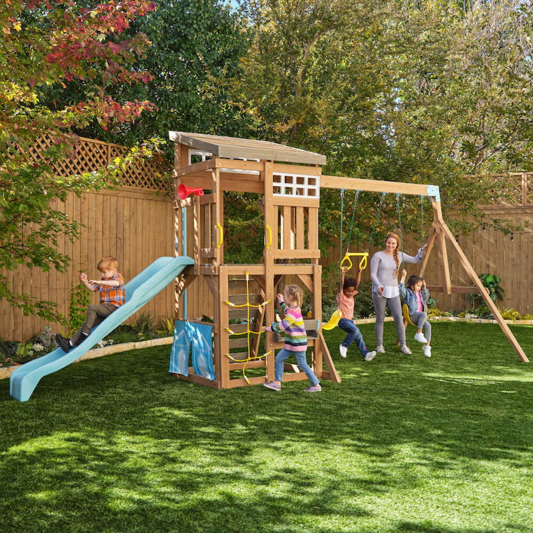 A KidKraft Modern Outdoor Wooden Swing Set sits in a grassy backyard surrounded by a wooden trellis fence and leafy trees with a group of children and woman playing in and around it as featured in Outdoor Home Playground Sets That Are Worth the Money for HGTV.ca