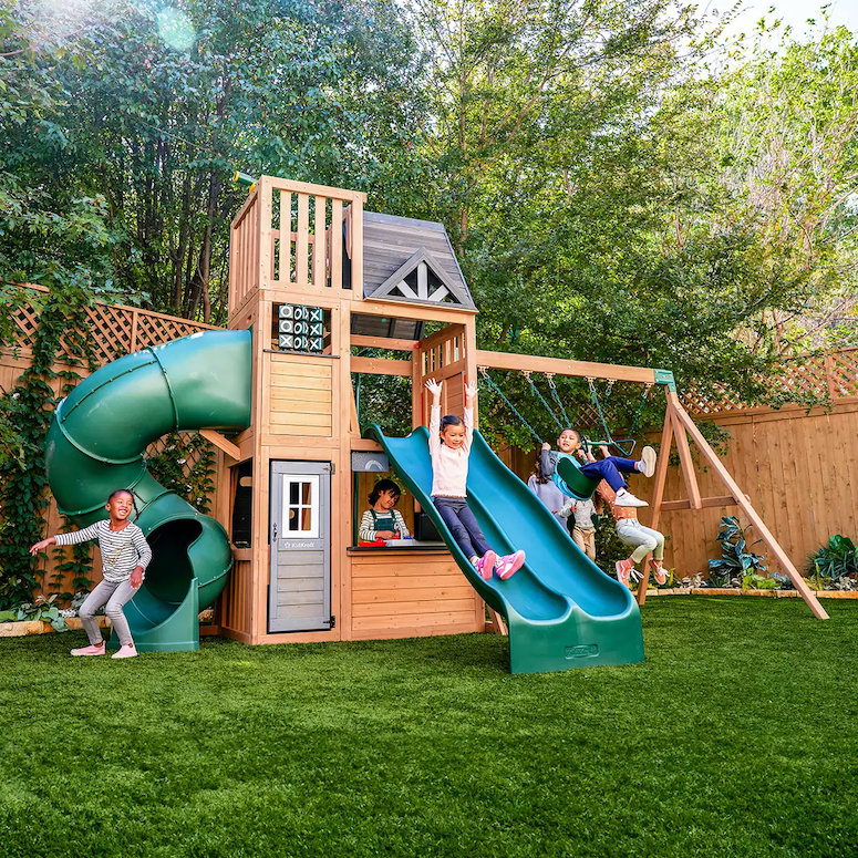 KidKraft Summit View Playset sits in a fenced backyard on a green lawn surrounded by trees with a group of kids playing on it as featured in Outdoor Home Playground Sets That Are Worth the Money for HGTV.ca