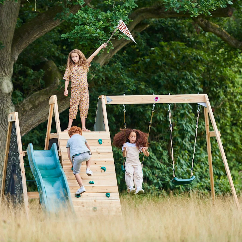Plum Play Residential Wood Climbing Pyramid Playset sits in a yellow grassy field in front of a large oak tree with a child holding a flag on top and two other children playing on it as featured in Outdoor Home Playground Sets That Are Worth the Money for HGTV.ca