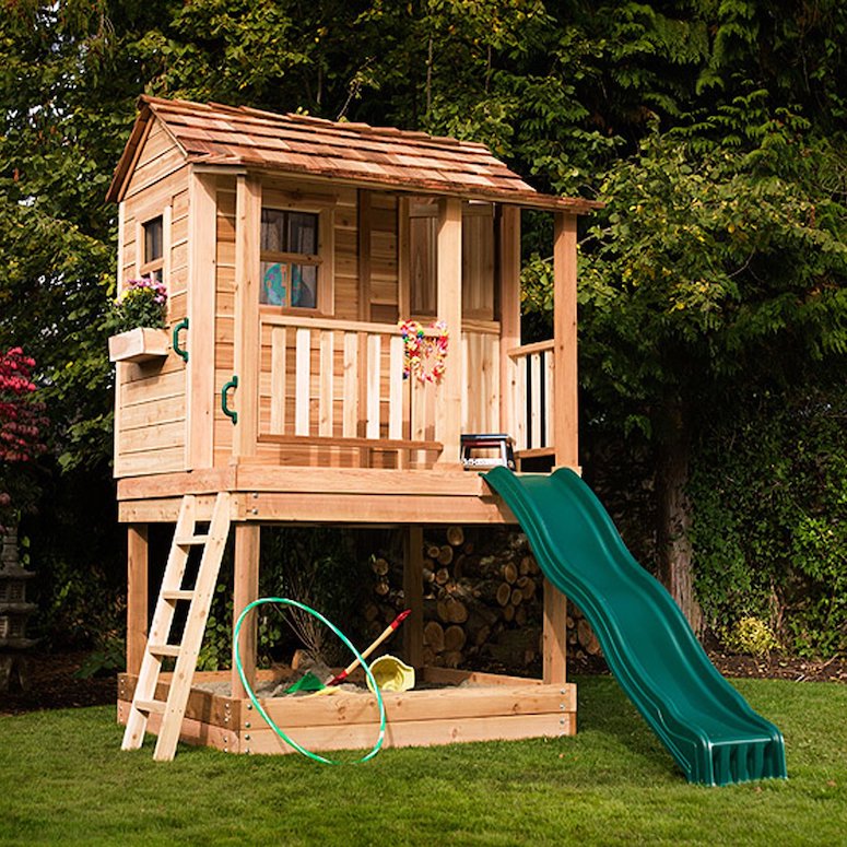 A sweet Little Cedar Playhouse & Sandbox by Outdoor Living Today features red cedar siding, a green slide, front porch and flower box and sits on green grass in front of a wooded area