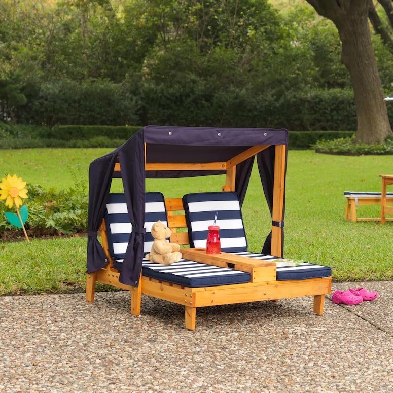 A navy and white Wooden Outdoor Double Chaise Lounge by KidKraft with a teddy bear sitting on it and a red water bottle in the bottle holder sits on a pebble rock patio next to a green lawn surrounded by a low hedge and trees as featured in Outdoor Home Playground Sets That Are Worth the Money for HGTV.ca