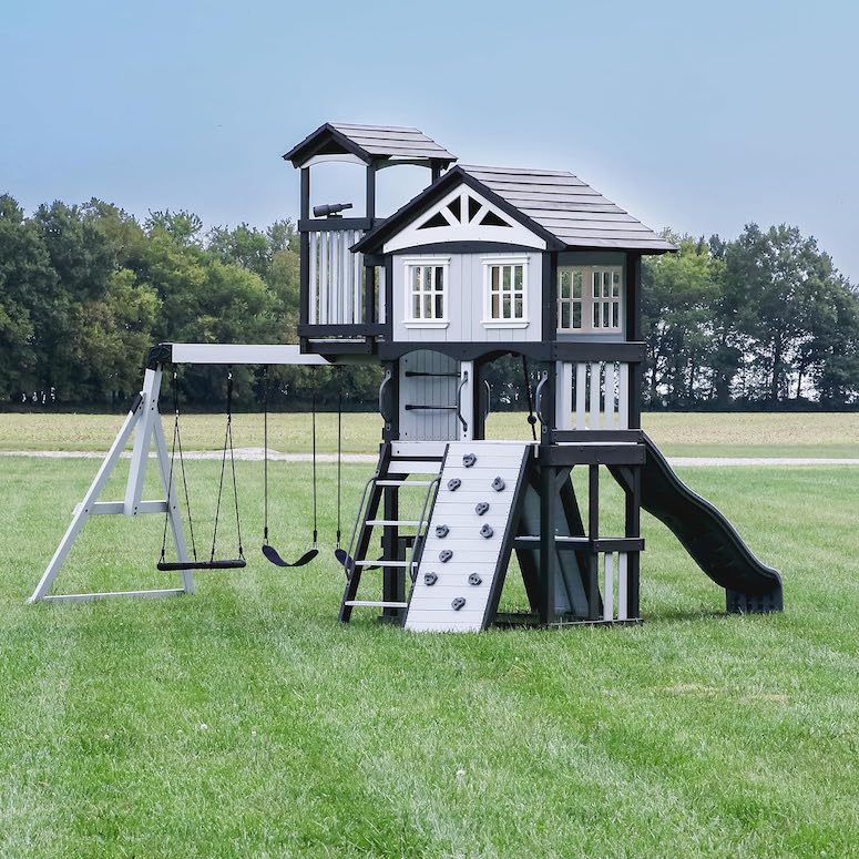 A large grey and black Whispering Point All Cedar Wooden Swing Set by Backyard Discovery sits in a large grassed field with trees in the background as featured in Outdoor Home Playground Sets That Are Worth the Money for HGTV.ca