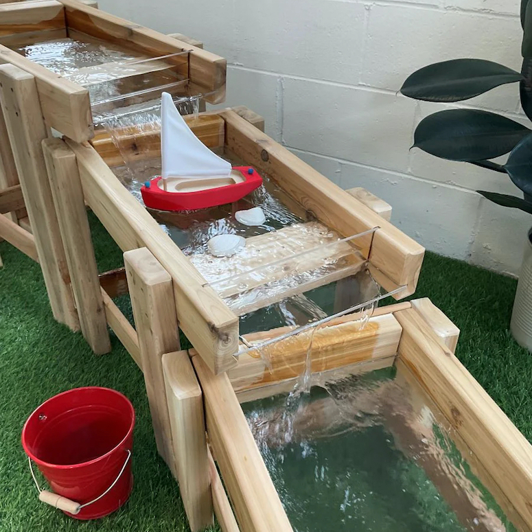Just Playing Cedar Water Flow Station sit on artificial turf in front of a white brick wall with a red wood boat flowing on the rushing water and red bucket on the ground as featured in Outdoor Home Playground Sets That Are Worth the Money for HGTV.ca