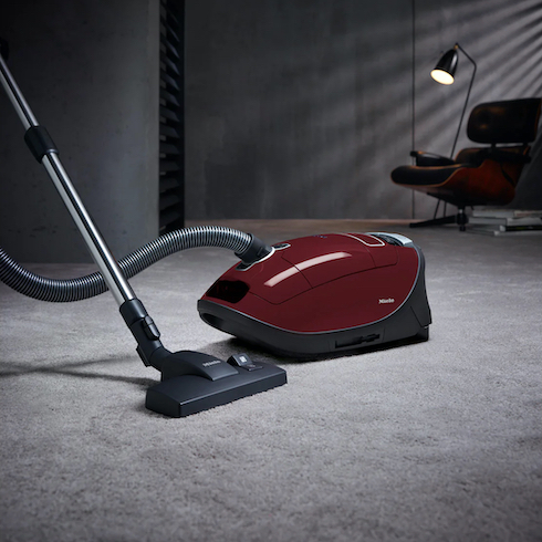 A Miele Complete C3 Limited Edition Cannister Vacuum sits on a grey carpet in a darkened room with an Eames armchair and lit floor lamp as part of How to Get Rid of Dust From Every Type of Surface at Home for HGTV.ca