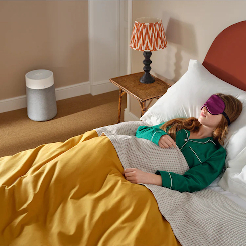 A Blueair Blue Pure 311i Max air filter sits on the floor next to a woman in green pyjamas and a purple sleeping mask asleep in a bed with yellow cover, white sheets, dark orange headboard and a bedside table with an orange and white lamp as part of How to Get Rid of Dust From Every Type of Surface at Home for HGTV.ca