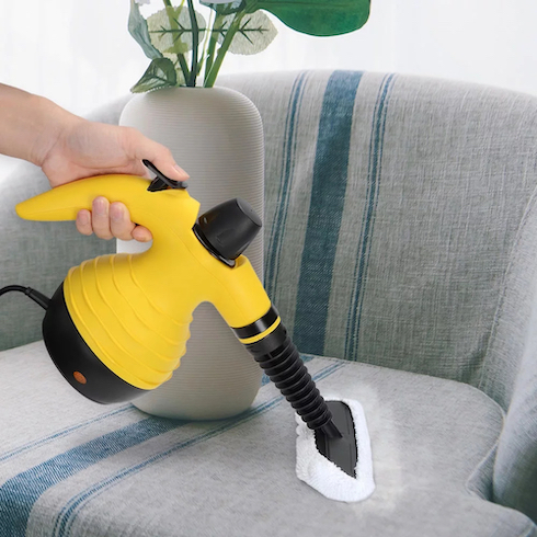 A hand as holds a yellow CleanWise All-In-One Handheld Steamer to steam a grey and grey armchair with a white vase holding faux greenery as part of How to Get Rid of Dust From Every Type of Surface at Home for HGTV.ca