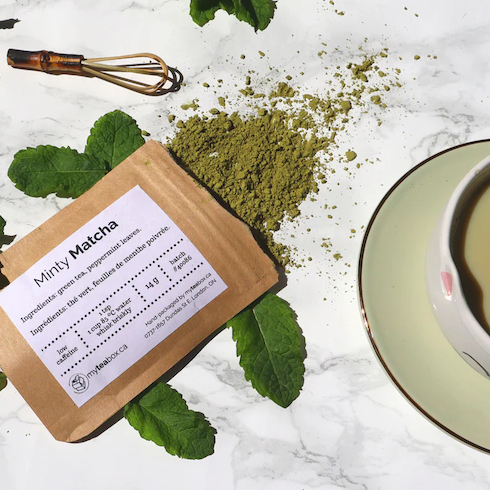 Packet of Minty Matcha from My Tea Box spills open onto a marble countertop with green mint leaves, a matcha whisk and a green cup and saucer as part of HGTV's 10 Gift Ideas for the Women in Your Life