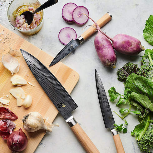 Set of Shun blonde handled knives on a grey countertop with a chopping board, garlic, onions, radishes, and green parsley as part of HGTV's 10 Gift Ideas for the Women in Your Life