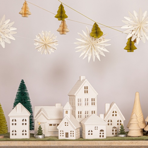 An array of white Christmas village decorations on table, under hanging paper Christmas trees and stars.