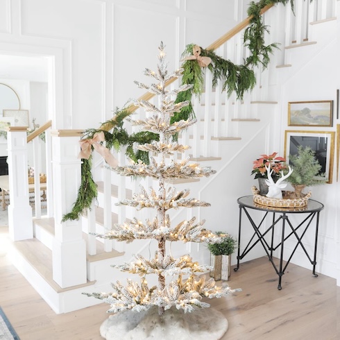 A white-flocked minimalist Christmas tree ists in front of a white staircase that's covered with green garland in a home entryway.