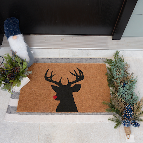 A silhouette of a red-nosed reindeer sits on a brown welcome mat outside a black front door of a home.