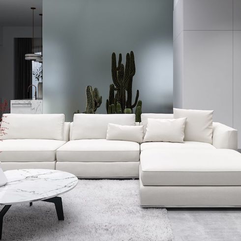 Cloud couch dupes featuring Rove Concepts Milo Modular Sectional in a low lit modern living area with a grey shag on a grey wood floor, a round marble topped coffee table and a large potted cactus in the background