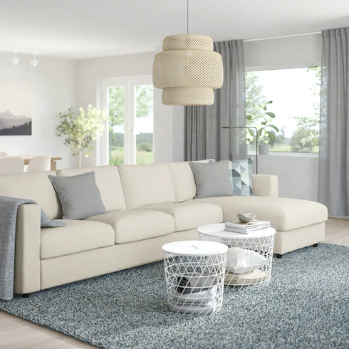 Cloud couch dupes featuring Ikea FINNALA sofa with a chaise lounge in Gunnared beige in a large living and dining area with light wood floors, large grass pendant lamp, a grey area rug, two white metal basket coffee tables, white walls, and grey gauzy drapes