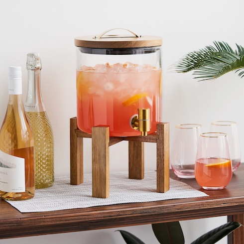 A wood-and-glass drink dispenser is filled with a festive orange beverage on a table.