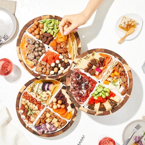 A hand reaches over three round trays that are each filled with an array of nuts, cheeses, meats, and snacks.